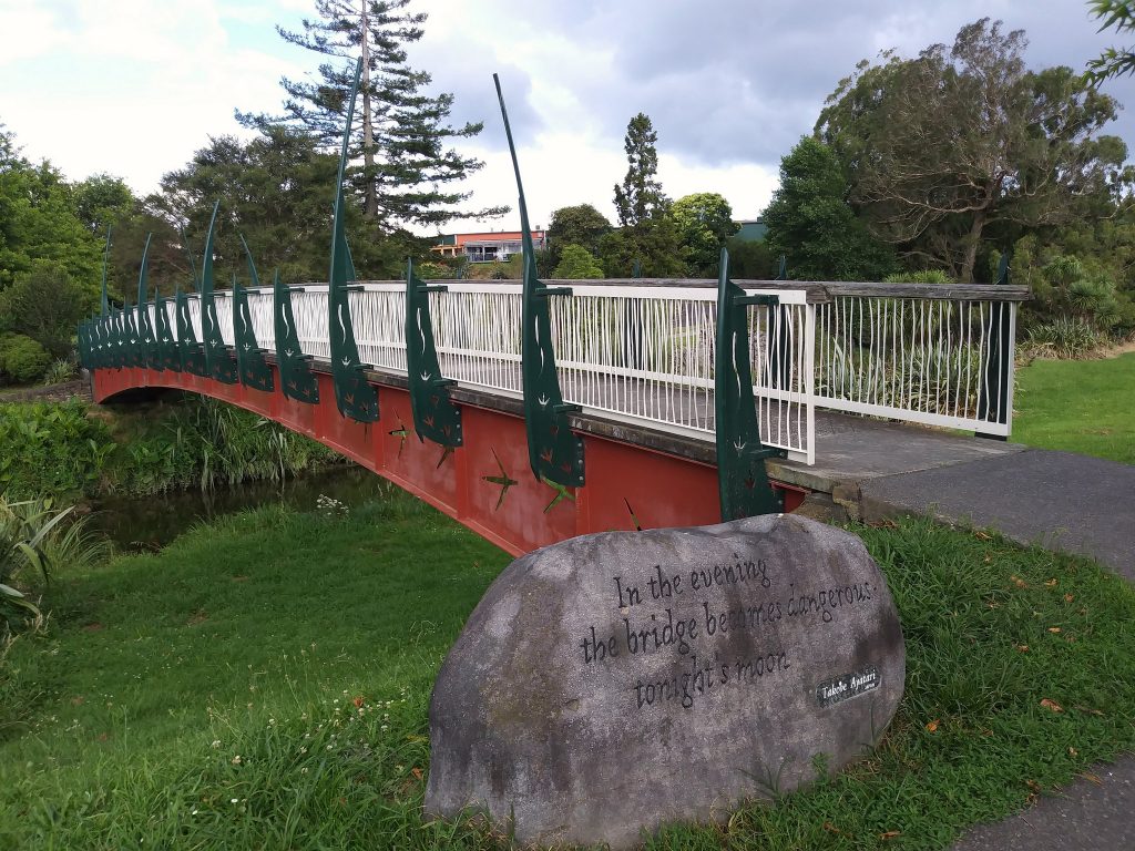 Red bridge with white rails and green decorative posts, across a stream. In front, a rock with a haiku.