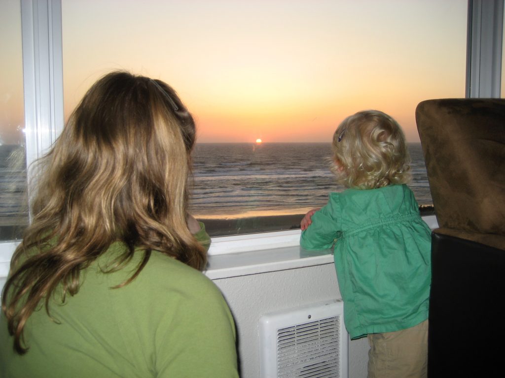 A woman and a toddler watch the sun set into the ocean outside a window